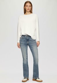 Witte lange mouw top met een ontspannen pasvorm, gecombineerd met lichtblauwe flare jeans en crèmekleurige schoenen met donkere veters en bruine zolen.