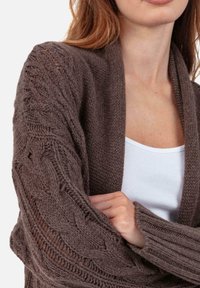 Cardigan marron tricoté avec motif torsadé, devant ouvert, poignets côtelés et manches texturées. Porté sur un débardeur blanc.