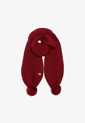 Écharpe rouge tricotée avec une texture côtelée épaisse, des pompons à chaque extrémité et une petite étiquette de marque rectangulaire.