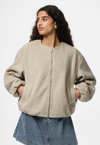 Jonge vrouw met lang krullend haar, gekleed in een beige zip-up jack en een blauwe spijkerrok, met haar handen in de zakken, kijkend naar links.