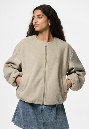 Jonge vrouw met lang krullend haar, gekleed in een beige zip-up jack en een blauwe spijkerrok, met haar handen in de zakken, kijkend naar links.
