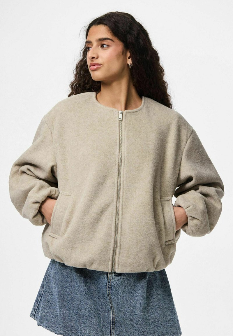 Jonge vrouw met lang krullend haar, gekleed in een beige zip-up jack en een blauwe spijkerrok, met haar handen in de zakken, kijkend naar links.