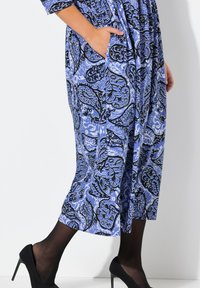 Largo vestido azul con estampado de paisley negro, con bolsillos laterales y ajuste suelto, combinado con zapatos de tacón negro.