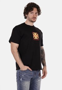 T-shirt in cotone nero con stampa grafica in rosso e giallo che raffigura un design stilizzato con la scritta "PROPAGANDA." Maniche corte.