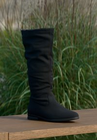 Botte noire montante en tissu doux avec un design froncé, bout arrondi et semelle en caoutchouc plate, présentée sur une surface en bois.