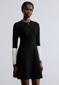 Čierne šaty so zapínaním na zips pri krku, s dlhými rukávmi, ktoré prechádzajú do bielej farby na manžetách. Hladká textúra a priliehavý strih.