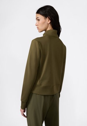 Vrouw met donker haar in een olijfgroene lange mouwen jas en bijpassende broek, zijwaarts kijkend tegen een effen witte achtergrond.