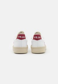 Sneaker bianche con suola beige, dotate di un tallone borgogna etichettato "VEJA" in testo bianco. Texture liscia con design minimalista.
