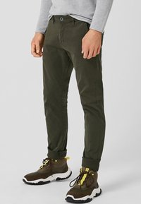 Chinos vert olive avec une coupe slim, en coton. Manches retroussées et fermeture à bouton. Portés avec des baskets marron et jaune.