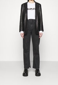 Schwarze Lederblazer über einem weißen T-Shirt mit "Levi's"-Logo, kombiniert mit hoch taillierten, dunkelblauen Jeans und schwarzen Plateau-Stiefeln.