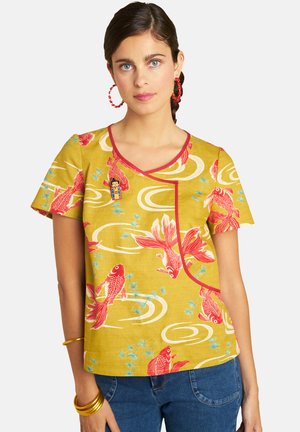 Woman wearing a yellow short-sleeve top with red koi fish and swirl patterns, paired with blue jeans and red hoop earrings.
