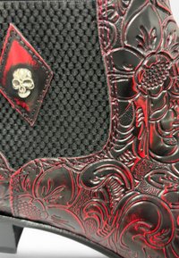 Bota negra texturizada con un patrón floral en relieve, acentos rojos y un detalle de calavera plateada. Suela resistente y un diseño distintivo.