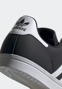 Czarno-biały sneaker Adidas pokazujący tylną piętę z czarnym logo trefoil na białej skórzanej łatce i teksturowanej białej podeszwie.