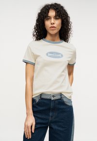 Κρεμ κοντομάνικη μπλούζα με μπλε λεπτομέρειες, με κυκλικό λογότυπο στο κέντρο. Συνδυάζεται με σκούρο τζιν παντελόνι.