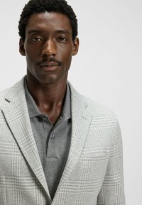 Jasnoszary żakiet w kratę założony na ciemnoszary polo. Żakiet ma klapy w stylu notched i jedną kieszeń na piersi. Gładka faktura.