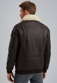 Brun læder bomberjakke med en cremet shearling-krave, ribbet kant og syninger, der har et mærke på venstre skulder.