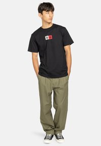 Zwart katoen T-shirt met een rechthoekig wit en rood grafisch ontwerp op de voorkant. Gecombineerd met losse olijfgroene broeken en zwarte sneakers.
