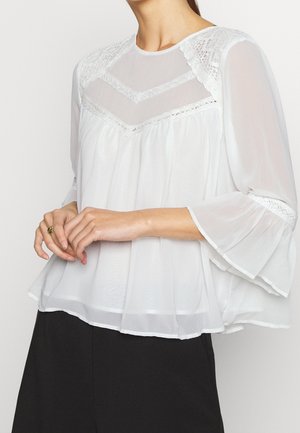 Kobieta ubrana w białą prześwitującą bluzkę z koronkowymi detalami na piersiach i dzwonowatymi rękawami, połączoną z czarnymi spodniami, ręce delikatnie złożone.