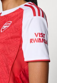 Maillot de sport rouge et blanc avec un design texturé rouge, col rond et le logo "VISIT RWANDA" en rouge en gras sur la manche.
