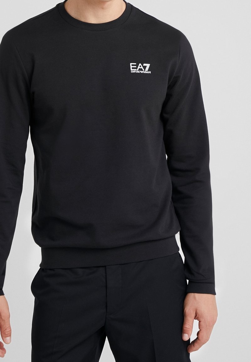 Homme portant un t-shirt noir à manches longues EA7 Emporio Armani et un pantalon noir, debout devant un fond clair uni.