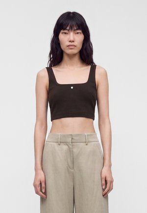 Jeune femme aux longs cheveux foncés portant un débardeur court noir et un pantalon beige taille haute, face à un fond uni.