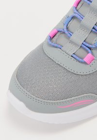 Zapatilla deportiva gris con un upper de malla texturizada, acentos en rosa, cordones azules y una suela de goma blanca. Destaca por un notable detalle de costura curva.