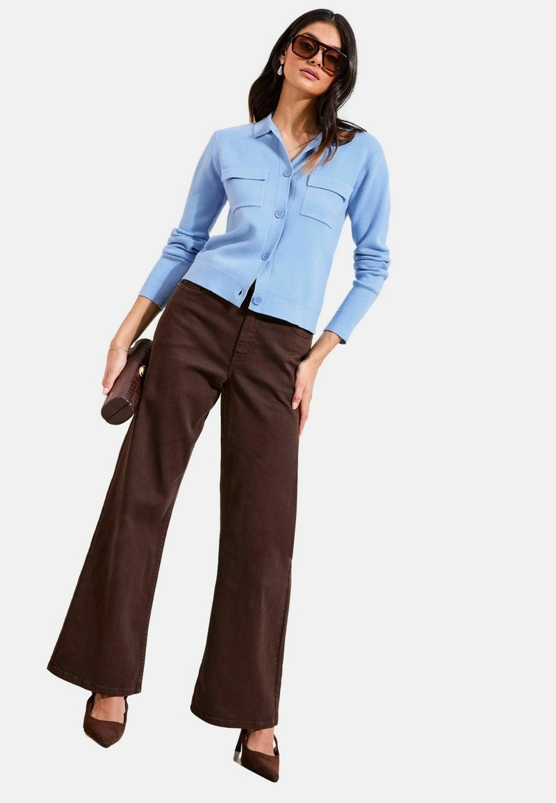 Frau trägt eine hellblaue, geknöpfte Strickjacke, braune Hosen mit weitem Bein, braune High Heels, Sonnenbrille und hält eine braune Clutch-Tasche.