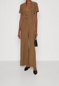Brun linne kortärmad jumpsuit med V-ringning, två knappar och vida ben, tillsammans med en liten svart handväska och svarta spetsiga skor.