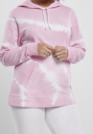 Persona con una sudadera con capucha en estilo tie-dye rosa y blanco con un bolsillo frontal, cordones blancos y pantalones blancos, manos entrelazadas con anillos en los dedos.