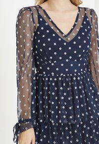 Námořnické šaty s průsvitnými dlouhými rukávy, vzorem puntíků, výstřihem do V, řaseným živůtkem a vrstvenou sukní s zavázanými manžetami.