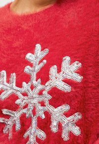 Pull rouge en peluche avec un grand motif de flocon de neige en sequins argentés, doté de branches détaillées et d'accents scintillants.