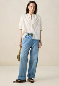 White short-sleeved blouse with decorative embroidery, light blue wide-leg denim pants, black sandals, and a small olive green handbag.