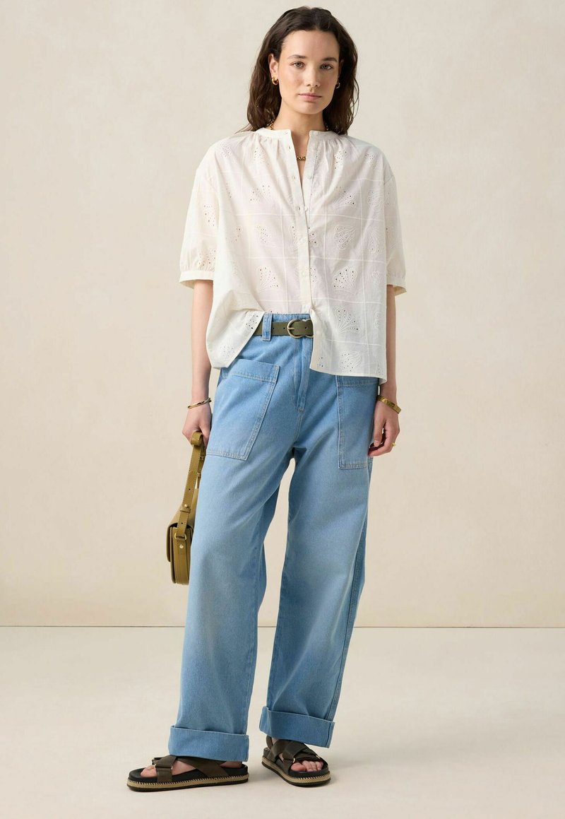 White short-sleeved blouse with decorative embroidery, light blue wide-leg denim pants, black sandals, and a small olive green handbag.