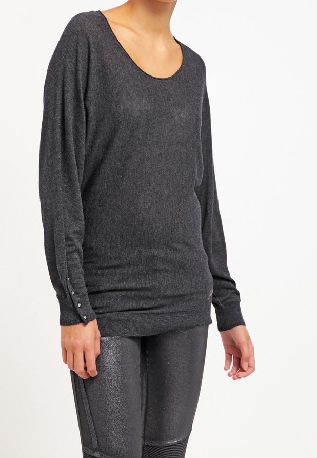 Femme portant un pull gris foncé à manches longues avec des détails de boutons sur les manches et des leggings noirs texturés brillants, debout devant un fond blanc.