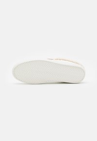 Sohle eines weißen Sneakers mit einem Fischgrätprofil, auf einer weißen Oberfläche ruhend, teilweise mit beigem, flauschigem Futter und goldenen Sternen-Details zu sehen.