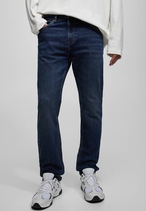 Osoba ubrana w ciemnoniebieskie dżinsy, białą koszulę z długim rękawem i białe trampki, stojąca z jedną ręką w kieszeni.