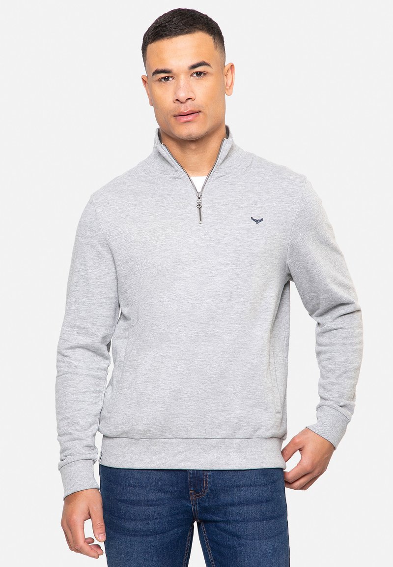 Sweat-shirt gris à col zippé en tissu doux, avec un ourlet et des poignets côtelés, et un petit logo sur la poitrine. Porté avec un jean bleu.