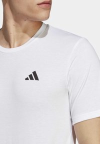 Detailní záběr na osobu, která má na sobě bílé tričko Adidas s černým logem Adidas na hrudi, obrácenou mírně doleva.