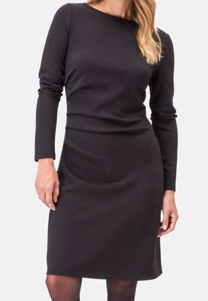 Frau trägt ein eng anliegendes, langärmeliges schwarzes Kleid mit U-Boot-Ausschnitt vor einem schlichten hellen Hintergrund.