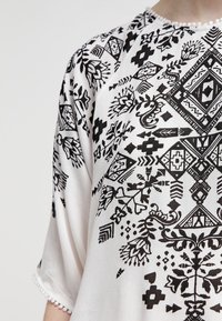 Blouse blanche ornée de motifs géométriques et floraux noirs complexes, manches courtes, et détails délicats en perles le long de l'ourlet.