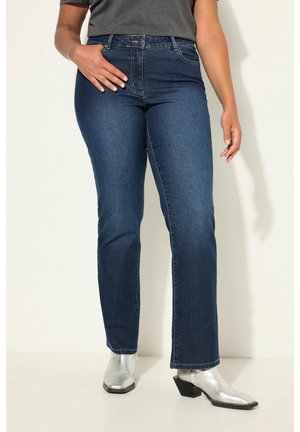 Persona che indossa jeans blu scuro a gamba dritta, maglia grigia e stivaletti metallizzati color argento, in piedi contro uno sfondo chiaro uniforme.