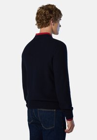 Suéter de punto azul marino con cuello redondo, acento rojo en el cuello, puños acanalados y dobladillo. Llevado con jeans de mezclilla oscura.