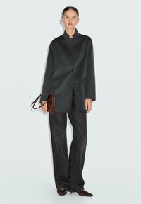 Cappotto di lana grigio scuro con un colletto alto e due tasche frontali, abbinato a pantaloni neri a gamba larga e una pochette in pelle marrone.