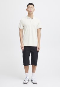 Svijetlosmeđa polo majica s kratkim rukavima, uparen s tamnim šortsevima do koljena. Bijele čarape i tenisice upotpunjuju ležerni outfit.