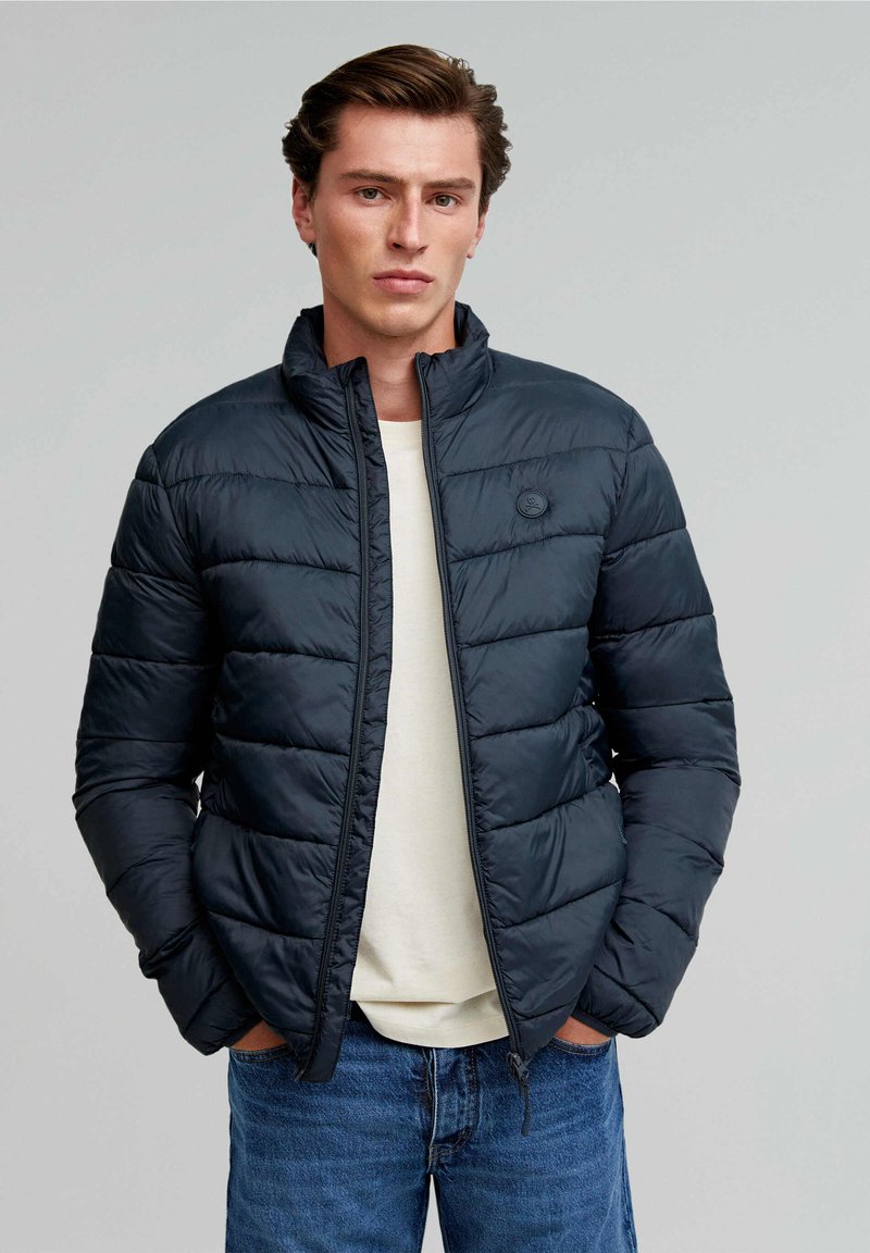 Veste rembourrée bleu marine foncé, avec un col montant et une fermeture éclair. Le tissu a une texture lisse avec un léger éclat.