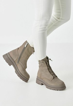 Personne portant des bottines taupe à lacets avec des fermetures éclair latérales et des semelles épaisses, associées à un pantalon blanc ajusté.