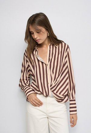Giovane donna con camicia beige e marrone a righe verticali con bottoni e maniche lunghe e pantaloni bianchi, in piedi davanti a uno sfondo semplice.