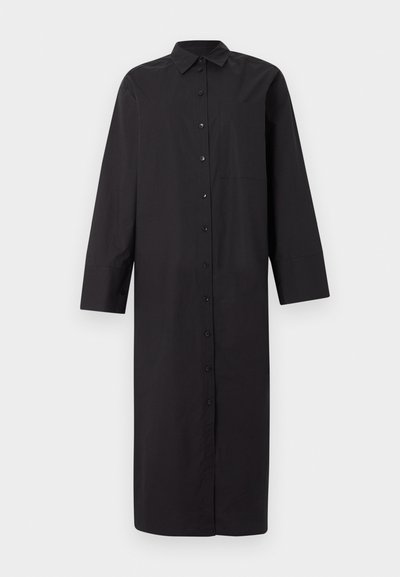 Μακρύ μαύρο φόρεμα πουκάμισο με μακριά μανίκια, γιακά και μπροστινή τσέπη, σε απλό ανοιχτό γκρι φόντο.