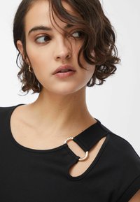 Haut noir avec un col arrondi, doté d'une bride accentuée avec un détail en anneau métallique. Texture lisse et manches courtes.