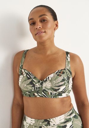 Kobieta stoi przy białej ścianie, ubrana w zielono-biały bikini w liście oraz dopasowane figi z wysokim stanem.